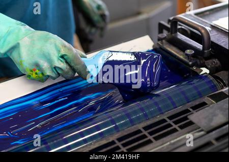 Par dessus l'employé anonyme dans des gants en caoutchouc sale étalant l'encre bleue sur les rouleaux de l'imprimante offset industrielle pendant le travail dans l'atelier de typographie Banque D'Images