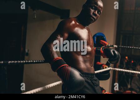 Un boxeur masculin en sueur regardant l'appareil photo tout en se reposant dans son coin de l'anneau de boxe. Jeune homme sportif ayant une séance d'entraînement dans un gymnase de boxe. Banque D'Images