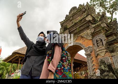 Deux musulmanes touristes femelles portant un foulard ou un hijab, prennent des selfies ensemble devant la porte du palais Ubud. Ils portent des masques pour éviter les covid- Banque D'Images