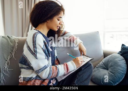 Belle femme sombre à la peau consultant réécrit l'information dans les papiers, études données, pose dans le cabinet spacieux à la maison, a une expression sérieuse. Prét Banque D'Images