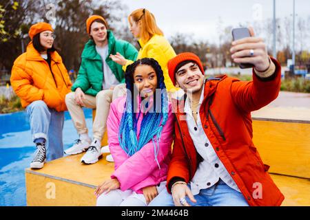 Groupe multiracial de jeunes amis heureux se rencontrant à l'extérieur en hiver, portant des vestes d'hiver et s'amusant, couple prenant le selfie sur smartphone pour soc Banque D'Images