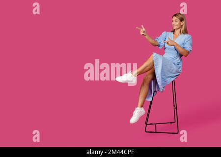 Bonne femme blanche blonde adulte dans la robe s'assoit sur la chaise, les doigts pointent à l'espace vide Banque D'Images