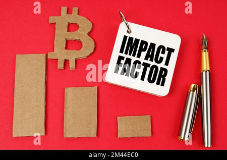 Concept Business et Bitcoin. Sur une surface rouge se trouvent un symbole de bitcoin, un graphique, un stylo et un bloc-notes avec l'inscription - FACTEUR D'IMPACT Banque D'Images