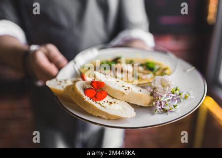 Serveur élégant servant un plat. Assiette avec pain et crevettes dans une sauce chaude. Arrière-plan flou. Concept de restaurant. Photo de haute qualité Banque D'Images