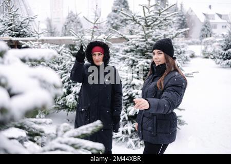 Fille dans la loi aider à acheter l'arbre de Noël pour la mère dans la loi en plein air à la foire. Enneigé Banque D'Images