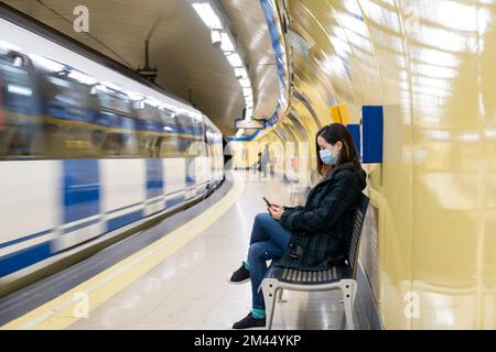 Jeune femme latine assise et portant un masque chirurgical avec un smartphone en attendant dans le métro ou la gare Banque D'Images