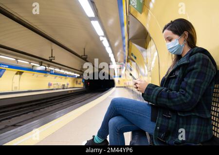 Jeune femme latine assise et portant un masque chirurgical avec un smartphone en attendant dans le métro ou la gare Banque D'Images