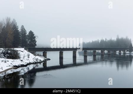 Un pont de chemin de fer traverse la paisible rivière Clark Fork par une journée d'hiver brumeuse en décembre dans le nord de l'Idaho. Banque D'Images