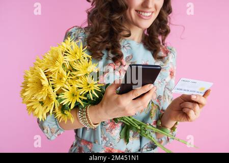 Gros plan sur Happy Modern Woman en robe florale avec fleurs de chrysanthèmes jaunes et carte de crédit à l'aide d'une application pour smartphone isolée sur fond rose. Banque D'Images