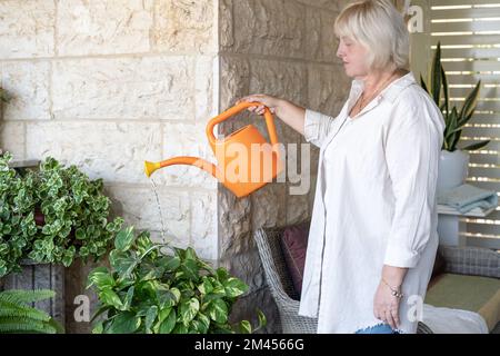 Belle heureuse femme d'âge moyen s'occupant du petit jardin local. Portrait d'une femme âgée souriante tenant un arrosoir et des fleurs arrosoir sur le patio. Banque D'Images
