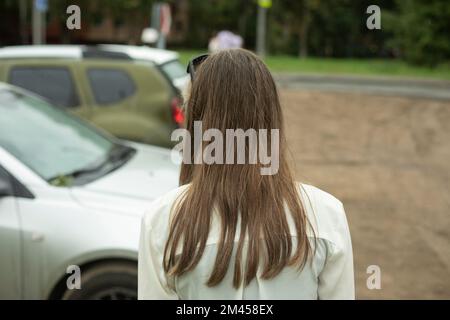 Fille dans le parking. Cheveux longs en été. Fille va à la voiture. Cheveux non colorés. Chemise blanche le jour de l'été. Banque D'Images