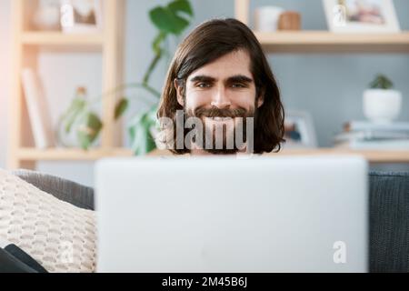 La connexion Internet est super et donne le sourire à tous. un beau jeune homme utilisant son ordinateur portable tout en se relaxant sur un canapé dans son salon. Banque D'Images