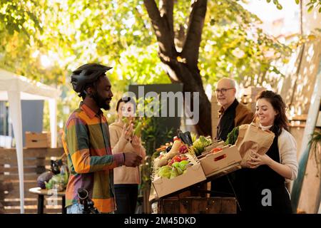 Un préposé local à la livraison d'aliments biologiques prend un sac de légumes pour donner l'ordre aux clients. Un couple d'agriculteurs locaux se tenant derrière un stand de fruits et légumes sur le marché des agriculteurs, donnant ordre au livreur. Banque D'Images