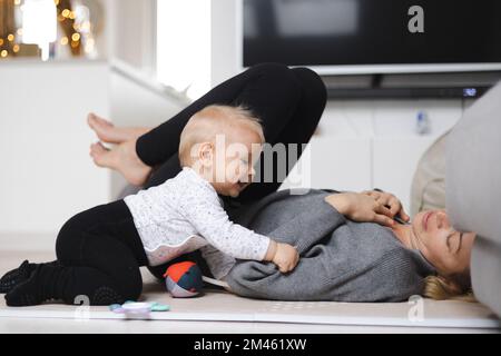 Des moments heureux en famille. Mère couchée confortablement sur le tapis des enfants jouant avec son bébé garçon en regardant et en défendant ses premiers pas. Émotions humaines positives, sentiments, joie Banque D'Images