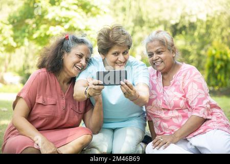 Des femmes indiennes heureuses utilisant un smartphone et prenant des photos d'elles-mêmes en plein air dans le parc d'été. Personnes âgées en cliquant sur selife . Banque D'Images