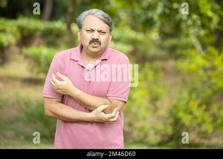 Homme indien âgé avec douleur au bras debout à l'extérieur du parc. Main d'homme ancien asiatique tenant son coude souffrant de douleurs au coude. Soins de santé. Banque D'Images