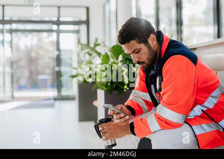 Le secouriste qui se brise, boit du café et fait défiler son téléphone portable dans le couloir de l'hôpital. Banque D'Images