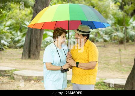 Joyeux couple indien senior debout sous un grand parapluie coloré portant des habits décontractés et un chapeau à l'extérieur du parc, vieux retraités appréciant des vacances. Banque D'Images