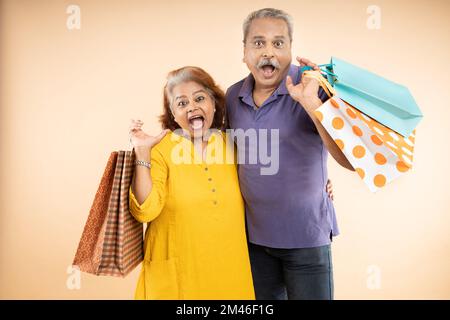 Un couple indien aîné surpris ou surpris portant des chiffons décontractés tenant des sacs de shopping ensemble isolés sur un fond beige. vie de retraite, e-commerc Banque D'Images