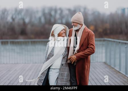 Élégant couple senior marchant près de la rivière, pendant la journée froide d'hiver. Banque D'Images