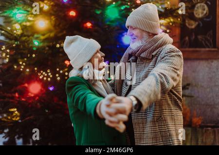 Couple senior ayant un moment romantique au marché de noël extérieur, dansant près de l'arbre de noël. Banque D'Images
