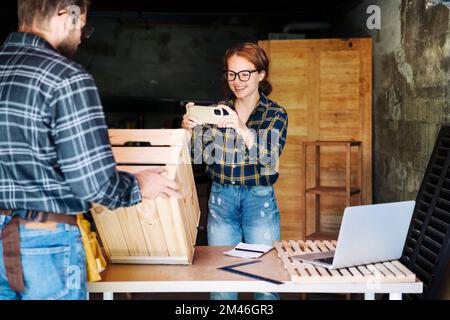 Jeune femme d'affaires prenant des photos de produits en bois à l'aide de son smartphone tout en travaillant dans son propre atelier. Banque D'Images