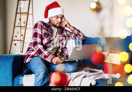 Seul homme en chapeau de père Noël assis à la maison et travaillant sur ordinateur portable pendant les vacances de Noël. Banque D'Images