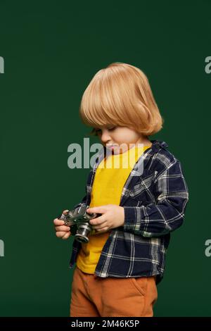 Portrait de petit garçon, enfant posant avec un appareil photo vintage sur fond vert studio. Photographe. Concept d'enfance, émotions Banque D'Images