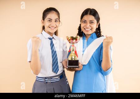 Happy Rural et moderne étudiant indien écolières portant l'uniforme de l'école tiennent le trophée de victoire dans la main debout ensemble isolé sur fond beige, W Banque D'Images