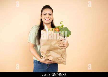 Heureuse belle femme indienne tenant un sac plein de légumes épicerie debout isolé sur fond beige. Gros plan Banque D'Images