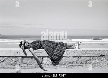Jeune française ou 1950s femme portant un Tartan, un motif à carreaux, une robe à carreaux ou à carreaux recrée ou se détend sur le Seawall à Hendaye Pyrénées-Atlantiques France 1950 Banque D'Images