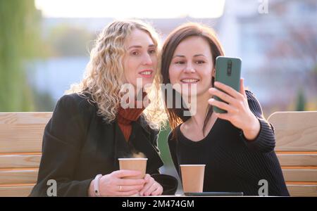 Deux femmes heureuses faisant le selfie avec un téléphone portable assis dans le café de rue de la ville. Des amies qui se réunissent à l'extérieur Banque D'Images