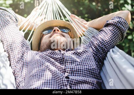 Homme barbu dans un chapeau et des lunettes de soleil couchés sur un hamac. Concept de vacances et de détente. Banque D'Images
