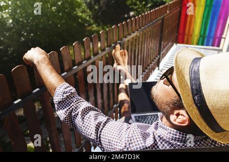 Homme en chapeau de paille et lunettes de soleil se détendant sur le balcon en été. Banque D'Images