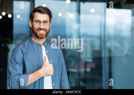 Portrait d'homme d'affaires dans une chemise décontractée, homme avec la barbe et les lunettes souriant et regardant l'appareil photo montrant les pouces affirmativement, propriétaire d'affaires travaillant à l'intérieur du bureau. Banque D'Images