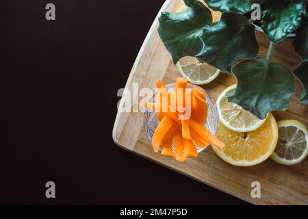 Vue de dessus d'un panneau en bois avec des tranches de citron, des bâtonnets de carottes et des feuilles vertes sur fond noir Banque D'Images