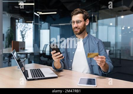 Homme d'affaires joyeux et souriant faisant des achats en ligne et le transfert d'argent de banque, indépendant tenant la carte de crédit de banque et smartphone, homme assis à un bureau à l'intérieur du bureau. Banque D'Images