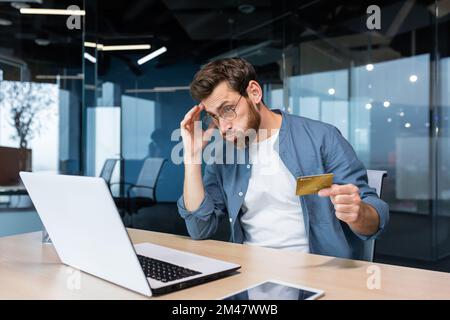 Triché et déçu homme d'affaires travaillant à l'intérieur du bureau, homme tenant la carte de crédit de banque et essayant de faire un achat dans la boutique en ligne, la fraude en ligne et la tromperie. Banque D'Images