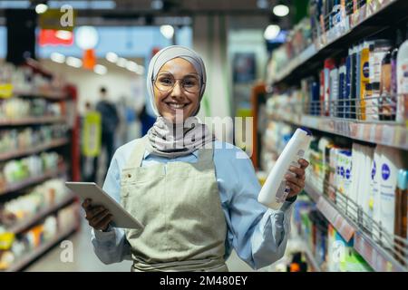 Portrait d'une femme de vente dans un hijab, un vendeur dans un département de produits chimiques domestiques sourit et regarde la caméra, tenant un ordinateur portable tablette dans ses mains. Banque D'Images