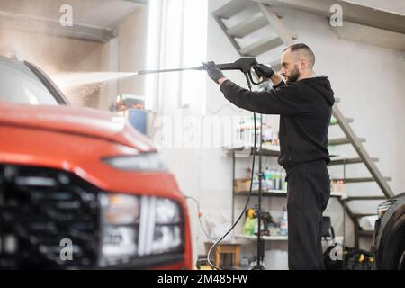 Portrait d'un jeune homme barbu qui lave la voiture rouge sous l'eau sous haute pression dans le service de lavage de voiture. Retirer le savon avec de l'eau, nettoyage de voiture. Banque D'Images