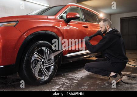 Roue, jante ou alliage de voiture, lavage. Nettoyage de voiture avec jet d'eau. Plan horizontal d'un jeune homme agréable en t-shirt blanc et combinaison, lavage de la roue rouge de voiture avec jet d'eau haute pression. Banque D'Images