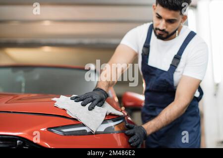 Vue à angle bas de l'ouvrier de lavage de voiture professionnel masculin dans des gants en caoutchouc noir, tenant la microfibre grise et polissage du capot de voiture et du phare de voiture rouge de luxe. Banque D'Images