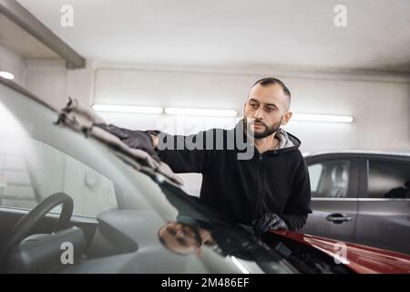 Concept de nettoyage et d'habillage de voiture. Un jeune homme de lavage de voiture de bonne qualité, portant des gants noirs, des lingettes et polit la fenêtre de pare-brise de voiture avec un chiffon en microfibre. Banque D'Images