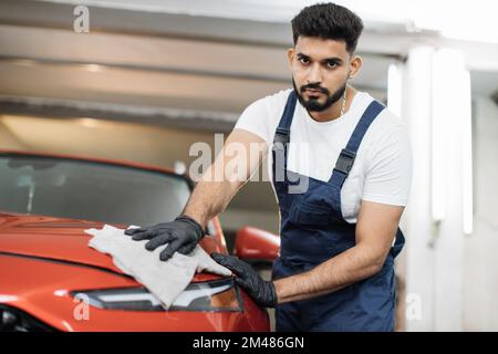 Vue à angle bas de l'ouvrier de lavage de voiture professionnel masculin dans des gants en caoutchouc noir, tenant la microfibre grise et polissage du capot de voiture et du phare de voiture rouge de luxe. Banque D'Images