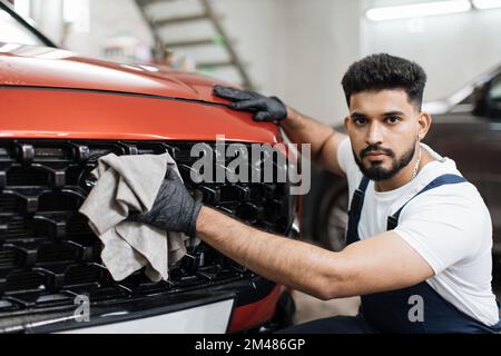 Concept de nettoyage, de lavage et d'habillage de voiture. Jeune homme barbu professionnel lave-auto dans des gants en caoutchouc noir, tenant la microfibre grise et la grille d'essuyage de voiture de luxe rouge. Banque D'Images