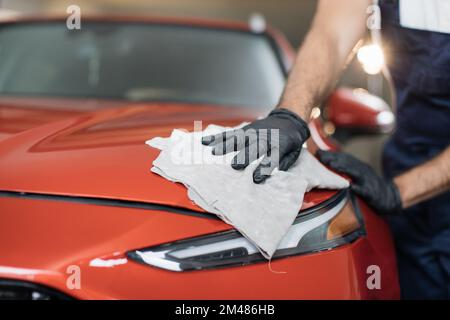 Image rognée de la main d'un ouvrier de lavage de voiture professionnel masculin dans des gants en caoutchouc noir, tenant la microfibre grise et polir le capot de voiture et le phare de voiture rouge de luxe. Banque D'Images