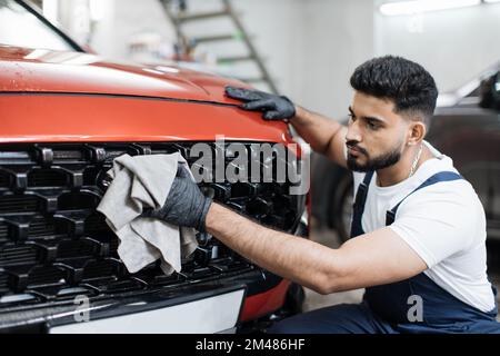 Concept de nettoyage, de lavage et d'habillage de voiture. Jeune homme barbu professionnel lave-auto dans des gants en caoutchouc noir, tenant la microfibre grise et la grille d'essuyage de voiture de luxe rouge. Banque D'Images