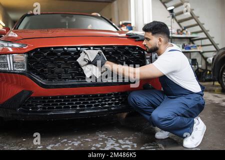 Vue latérale d'un ouvrier de lavage de voiture professionnel barbu dans des gants en caoutchouc noir, tenant la microfibre grise et la grille de radiateur essuyante de voiture rouge de luxe. Banque D'Images