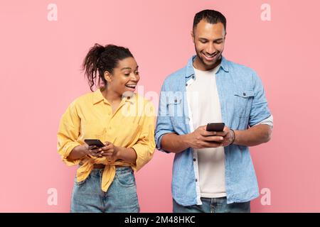 Curieuse femme noire essayant de regarder le téléphone portable de son petit ami, homme utilisant son gadget et de chat, fond rose Banque D'Images