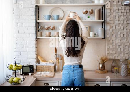 Jeune femme caucasienne mettant des assiettes et des bocaux sur étagère, debout dans la cuisine intérieure, vue arrière, espace libre Banque D'Images